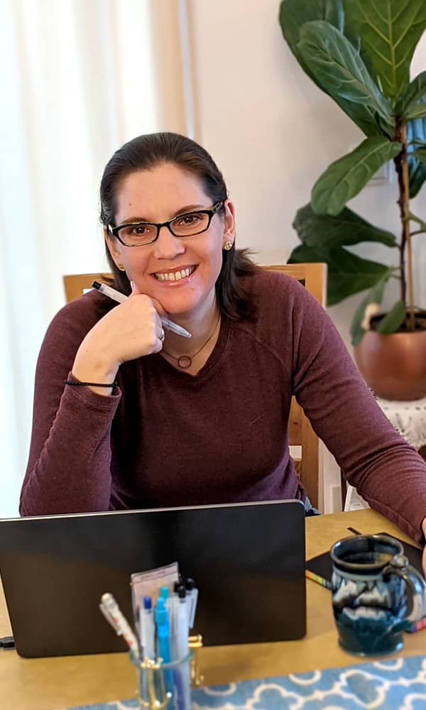 Stephanie sits at a work table behind her laptop. She wears glasses, a burgundy sweater, and a broad smile
